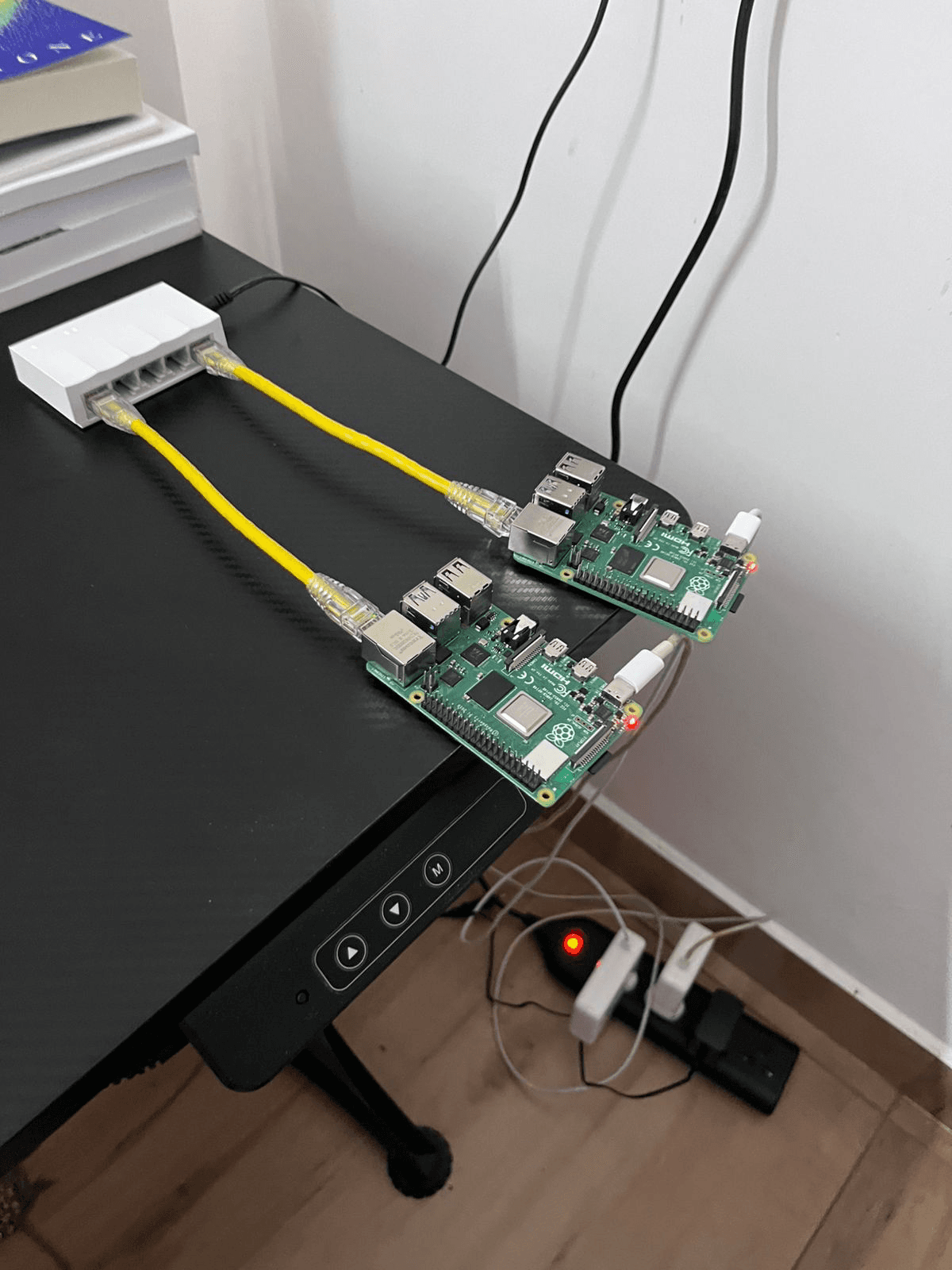 My Raspberry Pi cluster running Kubernetes - Two Raspberry Pi 4s connected to a small white desktop ethernet switch with short yellow ethernet cables. You can see two Macbook adapters connected powering the Pis, plugged into a power strip along with the power adapter of the ethernet switch